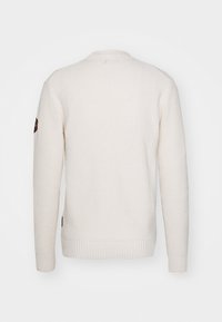 Waffelineulottu kermainen neulepaita, jossa on pyöreä kaula-aukko, resorit hihansuissa ja helmassa, vasemmassa hihassa merkki ja teksturoidut yksityiskohdat.