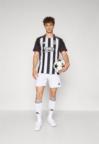 Mustvalge triibuline jalgpallimängija särk punaste detailidega, valged lühikesed püksid ja mustvalge jalgpall; modell kannab valgeid sokke ja kummikuid.