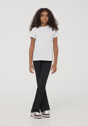 T-shirt bianca in cotone a maniche corte, abbinata a pantaloni neri a zampa d'elefante e sneakers bianche con dettagli bordeaux, in piedi davanti a uno sfondo semplice.