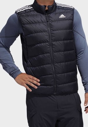 Mężczyzna w czarnej pikowanej kamizelce Adidas na niebieskiej sportowej koszulce z długim rękawem, stoi z jedną ręką na biodrze na jednolitym tle.