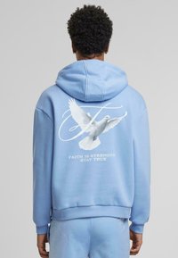 Felpa leggera blu chiaro con una grafica di una colomba bianca e il testo "LA FEDE È FORZA RIMANI VERITIERO" sul retro. Realizzata in un tessuto morbido e testurizzato.