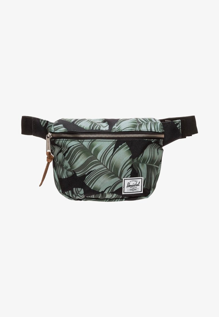 Sac banane avec motif feuilles tropicales noir et vert, poche zippée frontale, sangle noire réglable et étiquette de marque rectangulaire blanche.