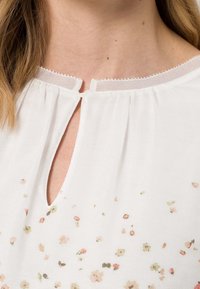 Blusa blanca con escote redondo, detalle en forma de llave y delicado estampado floral en colores pastel. Tela texturizada con caída suave.