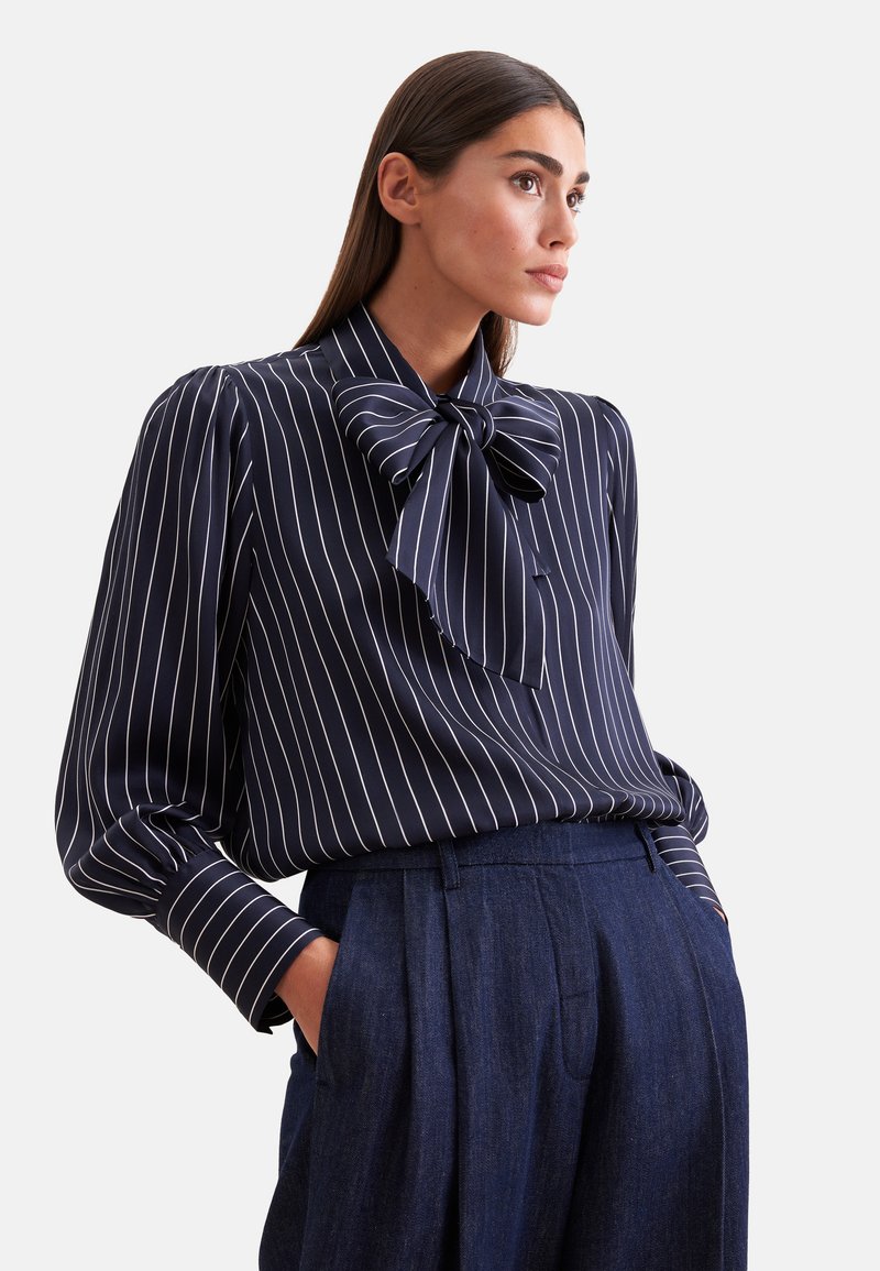 Femme aux longs cheveux foncés porte un chemisier bleu marine à fines rayures blanches et un grand nœud, associé à un pantalon large bleu foncé taille haute.