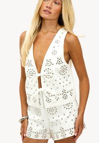 Mujer rubia vestida con un chaleco sin mangas blanco y pantalones cortos con patrones de tachuelas cuadradas plateadas y cierres delanteros con nudos, complementada con una pulsera plateada.