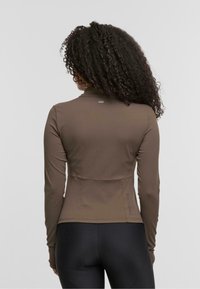 Langärmliges, braunes Sportoberteil aus elastischem Material, mit hohem Kragen und dezentem Branding auf der Rückseite. Getragen mit schwarzen Leggings.