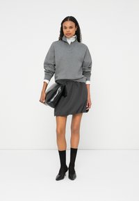 Γκρι πουλόβερ με γιακά, συνδυασμένο με μια σκοτεινή πλισέ φούστα. Το μοντέλο κρατάει μια μαύρη γυαλιστερή τσάντα. Μαύρες κάλτσες και μυτερές γόβες ολοκληρώνουν την εμφάνιση.