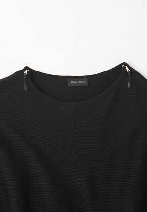 Sweater preto canelado com decote redondo, ligeira textura e detalhes em zíper prateado nos ombros. Etiqueta lê "ANNA FIELD."