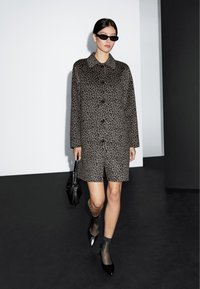 Cappotto in fantasia leopardata in tonalità grigie con colletto, bottoni frontali e maniche lunghe, abbinato a una borsa nera e scarpe a punta.