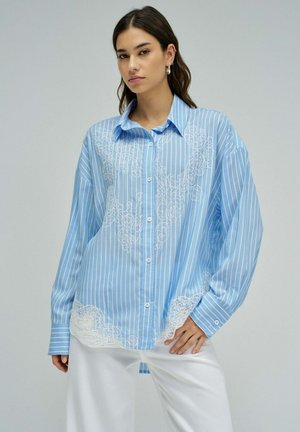 Jeune femme portant une chemise à boutons bleu clair rayée de blanc avec des détails en dentelle blanche, associée à un pantalon blanc ample, posant devant un fond uni.