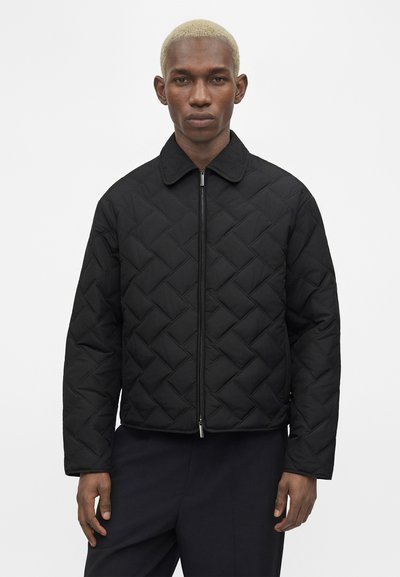 Άνδρας με κοντά ξανθά μαλλιά που φοράει μαύρο μπουφάν με φερμουάρ και σκουρόχρωμο παντελόνι, στέκεται μπροστά σε λευκό φόντο.