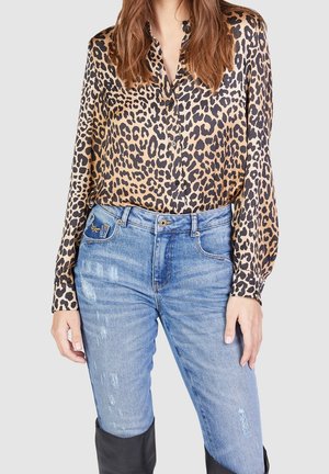 Frau trägt eine langärmelige Bluse mit Leopardenmuster, die in eine hellblaue, verwaschene Jeans gesteckt ist, und schwarze kniehohe Stiefel vor einfachem Hintergrund.