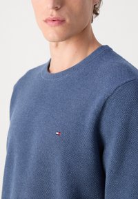 Blå strikket genser med et teksturert mønster, ribbet rund hals, og en liten brodert logo i rødt, hvitt og blått på brystet.