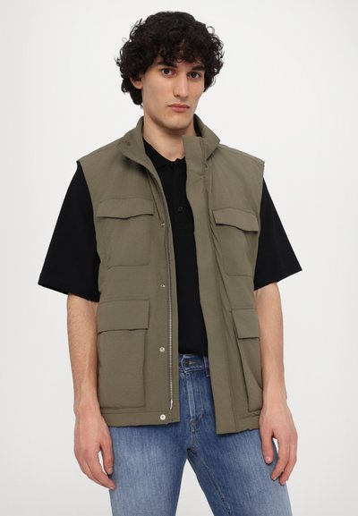 Olijfgroene mouwloze vest met een ritssluiting aan de voorkant, meerdere voorzakken en een relaxte pasvorm, gecombineerd met een zwart poloshirt en blauwe jeans.