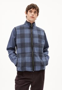 Männer Steppweste in Blau mit Karomuster, ausgestattet mit hohem Kragen, Seitentaschen und einem Logo auf der Brust. Hergestellt aus weichem Stoff.