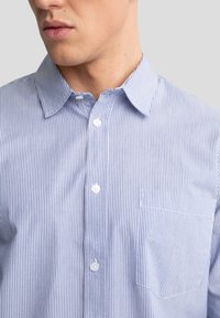 Camicia button-up a righe blu e bianche con colletto standard, dotata di taschino e texture del tessuto liscia. Bottoni bianchi lungo il davanti.
