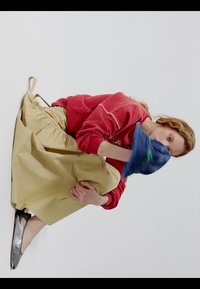 Donna con felpa rossa e gonna beige seduta rannicchiata, che tiene un cuscino blu contro il viso su sfondo bianco.