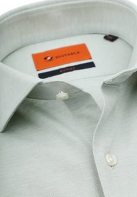 Lichtgroene, gebreide shirtkraag van een textuurstof, met een enkele knop en een oranje merkteken met zwarte accenten aan de binnenkant.