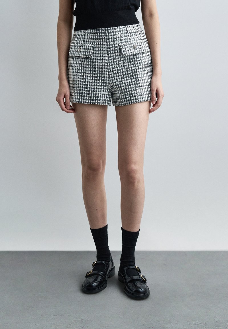 Γυναίκα που φοράει ψηλόμεσο σορτς με ασπρόμαυρο καρό, εμπρόσθιες τσέπες, μαύρες κάλτσες μέχρι τον αστράγαλο και μαύρα παπούτσια με αγκράφα, στέκεται σε γκρι πάτωμα.