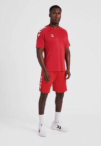 Rood sportuniform bestaande uit een shirt met korte mouwen en een korte broek. Heeft contrasterende witte accenten en een logo op het shirt. Draagt witte sneakers.
