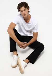 Camiseta de algodón blanca, pantalones negros y zapatillas de deporte de color claro. Modelo sentado con las rodillas levantadas, manos entrelazadas. Diseño minimalista, colores neutros.