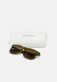 Ochelari de soare Calvin Klein Jeans cu un cadru translucid de culoare verde închis și lentile maro, așezați lângă o carcasă albă dură cu logo.