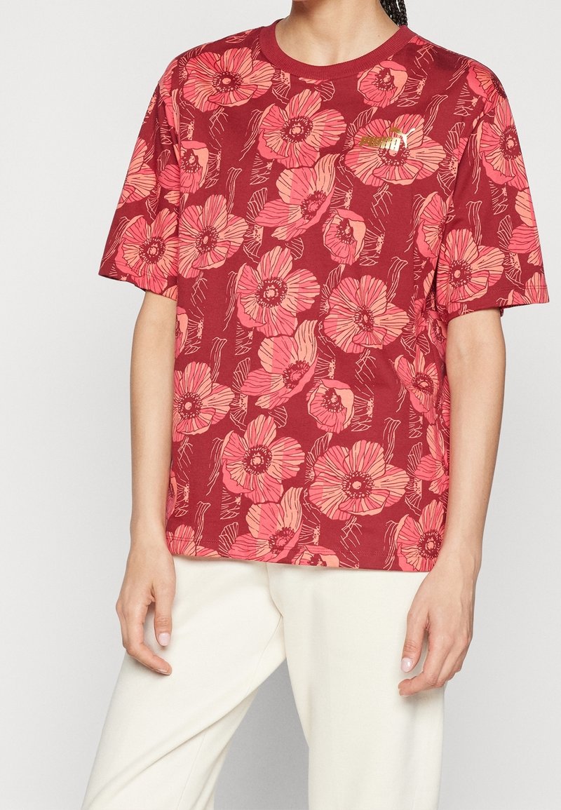 Personne portant une chemise rouge à manches courtes avec de grands motifs floraux roses et un pantalon blanc, montrée des épaules aux cuisses.