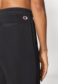 Μαύρο παντελόνι φόρμας με ελαστική μέση, πλαϊνές τσέπες και μικρό κεντητό λογότυπο. Λεία υφή και σχεδιασμός με στενή γραμμή στα πόδια.