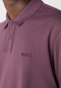 Gros plan d'une personne portant un polo couleur prune avec deux boutons et « BOSS » brodé sur la poitrine gauche.