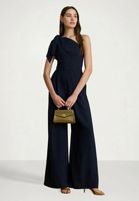 Žena stojí v námořní modré overalovej kombinéze s jedným ramenom a drží malú olivovo zelenú kabelku, na nohách má béžové lodičky.