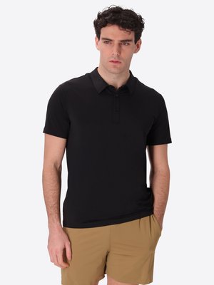 Giovane uomo con capelli ricci scuri indossa una polo nera a maniche corte e pantaloncini beige su uno sfondo bianco semplice.