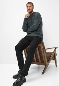 Maglione grigio scuro a maglia con motivo testurizzato, abbinato a pantaloni neri e scarpe nere. Il modello è seduto su una sedia di legno con un cuscino marrone.