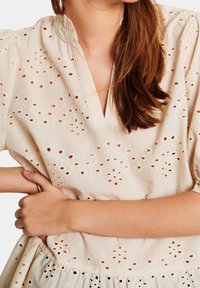 Ljust beige broderad topp med korta puffärmar, som har hålmönster och en v-ringning. Mjuk, strukturerad tyg.