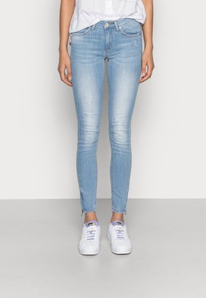 Jeans skinny de color azul claro, fabricados en denim, con un lavado desvaído, ligero desgaste y dobladillos con cremallera. Combinados con zapatillas blancas.