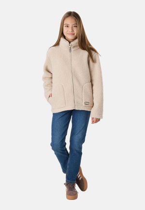 Ung person står med hænderne i lommerne iført en beige, blød lynlåstrøje, blå jeans og brune sneakers med hvide striber.