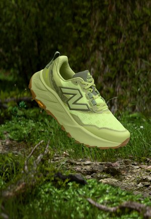 Scarpa da trail running New Balance verde chiaro con suola Vibram che fluttua sopra un fondo roccioso in una foresta con sfondo di fogliame verde.