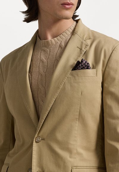 Beige bavlněný blazer se dvěma knoflíky, v kombinaci se světle béžovým svetr se vzorem pleteniny. Kapesní čtverec má tmavé pozadí s barevnými vzory.