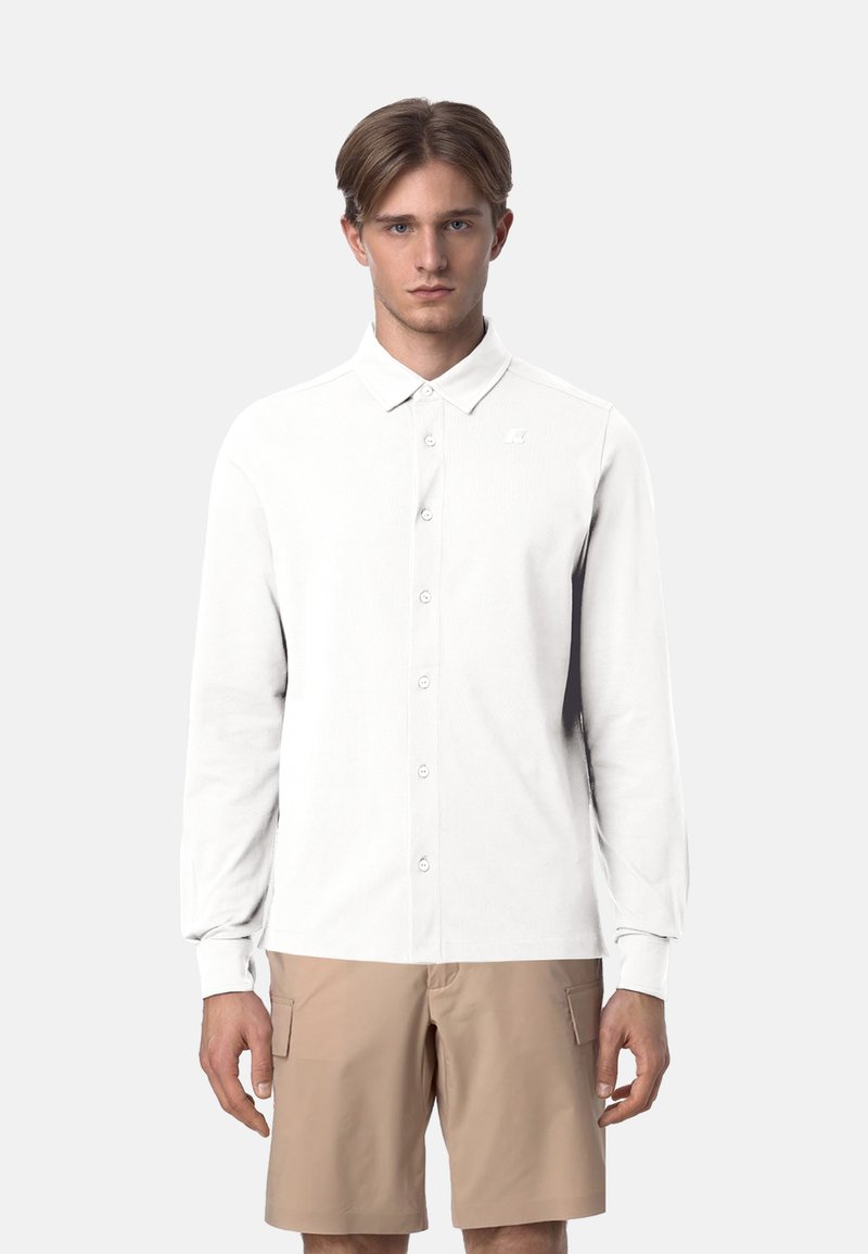 Polo bianco a maniche lunghe con colletto button-down, caratterizzato da un logo sottile sul petto e un tessuto liscio e leggero. Abbinato a pantaloni corti beige.