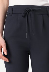 Marinblå byxor med dragsko i midjan, veck och en mjuk tygtextur. Inkluderar sidofickor och subtila sömdetaljer.