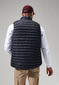 Marineblauwe gewatteerde vest met horizontaal gestikt ontwerp, heeft een hoge kraag en is gecombineerd met een wit langslav shirt en beige broek.