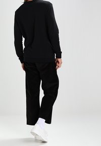 Person som bär en svart långärmad tröja, svarta kordbyxor, vita strumpor och vita sneakers, som står mot en enkel ljus bakgrund.