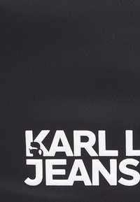 Material textil de culoare neagră, cu litere albe mari și proeminente care spun "KARL LAGERFELD JEANS", având o suprafață texturată și un design minimalist.