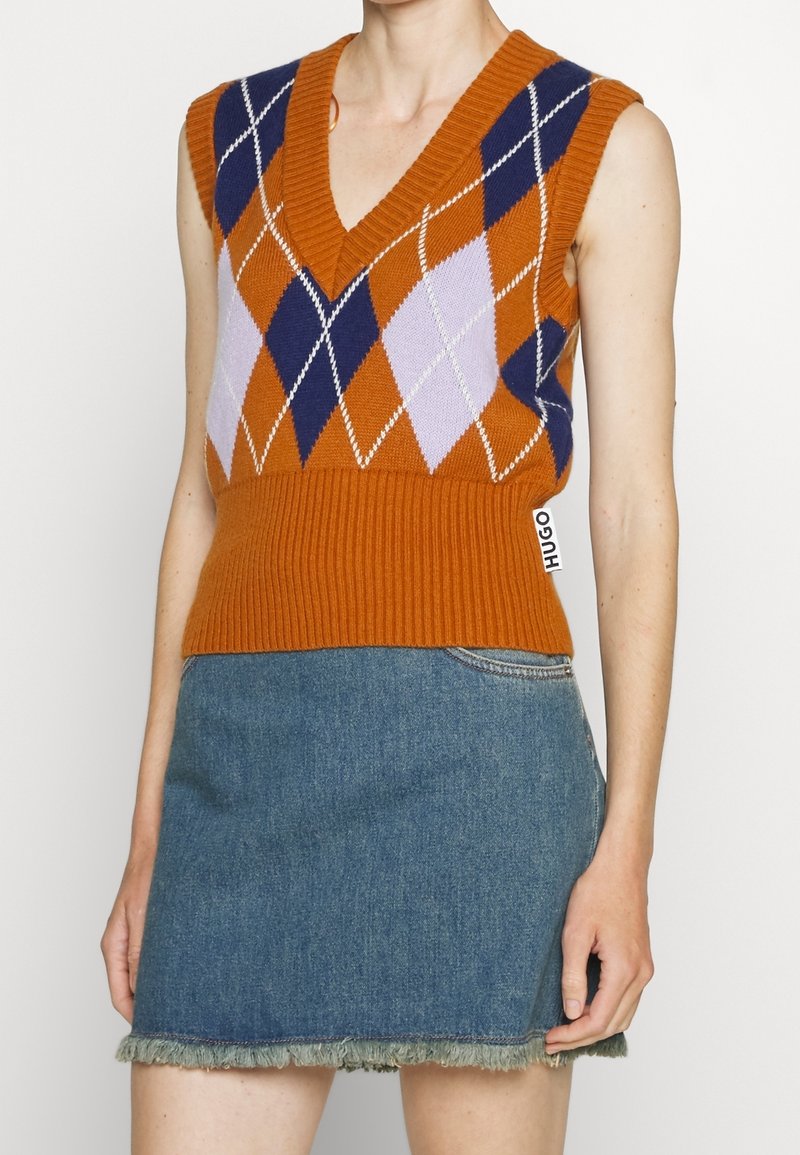 Veste tricotée sans manches orange à losanges bleus et lavande, portée avec une mini-jupe en jean bleu avec un ourlet effiloché.