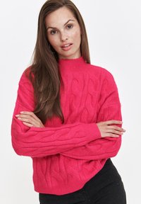 Jasnoróżowy sweter z dzianiny w warkocze z wysokim dekoltem i ściągaczami. Materiał wydaje się miękki i teksturowany, z wyrazistym wzorem.