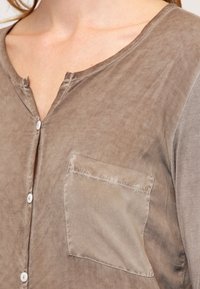 Camicia marrone con bottoni, dal tessuto morbido, con taschino sul petto e colletto arrotondato. Il tessuto ha una leggera lucentezza.