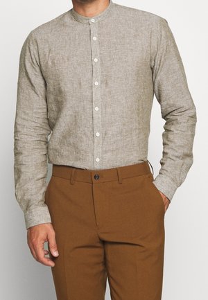 Ein Mann trägt ein hellbeiges Leinenhemd mit Mandarinkragen, das in braune, maßgeschneiderte Hosen gesteckt ist, und steht mit einer Hand in der Tasche.