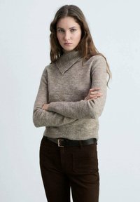 Beige, tætsiddende turtleneck sweater lavet af blødt materiale. Sammen med brune bukser og et sort bælte. Enkel tekstur, ingen synlige mønstre.