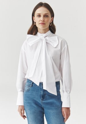 White blouse with a large bow at the collar, long sleeves, and a smooth texture, paired with light blue denim jeans.