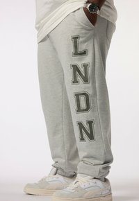 Grijze joggingbroek met vetgedrukt "LNDN"-tekst in donkergroen. Gemaakt van zachte stof, met een comfortabele pasvorm en zijzakken. Gecombineerd met lichtgekleurde schoenen.