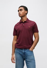 Bordowa koszulka polo z kołnierzykiem i krótkimi rękawami, z małym haftowanym logo na piersi, połączona z jasnoniebieskimi dżinsami.